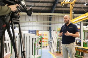 man stood in factory, in front of window frames, being recorded by a camera